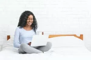 Beautiful smiling young woman sitting on bed, using laptop